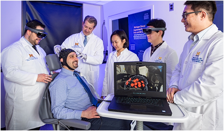 Hassan Jassar (seated) wears a sensor-outfitted cap that detects changes in blood flow and oxygenation, thus sensing brain activity. That information is transmitted to a computer and interpreted. Thiago Nascimento, left, views this brain activity in real-time while wearing augmented reality glasses, and the computer image shows that particular pain signature in the brain. From left to right, also pictured, Dr. Alex DaSilva, Dajung Kim, Manyoel Lim, Xiao-su Hu.